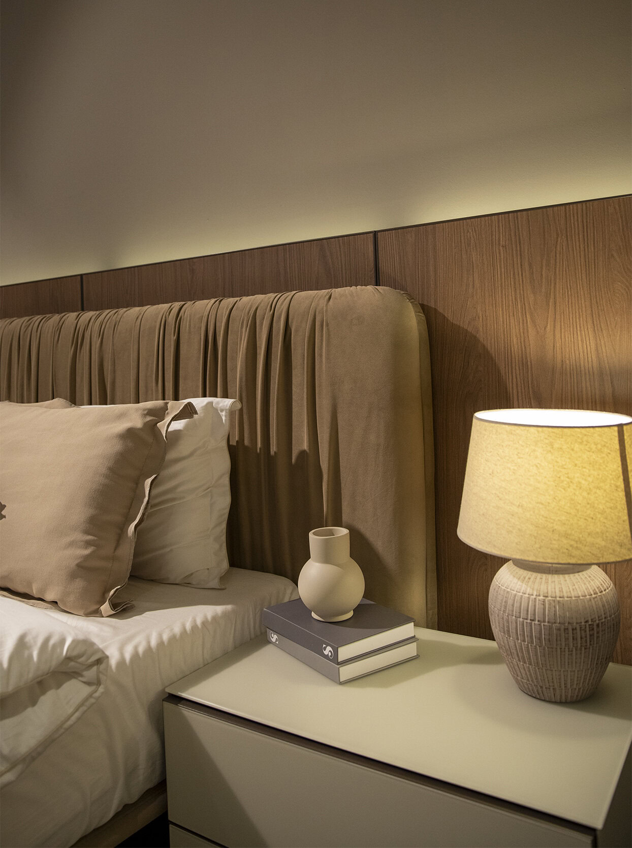 Elegant bedroom with a wooden headboard, beige bedding, and modern accessories including a table lamp, decorative vase, and stacked books, showcasing stylish and comfortable furniture from Saloni Furniture.