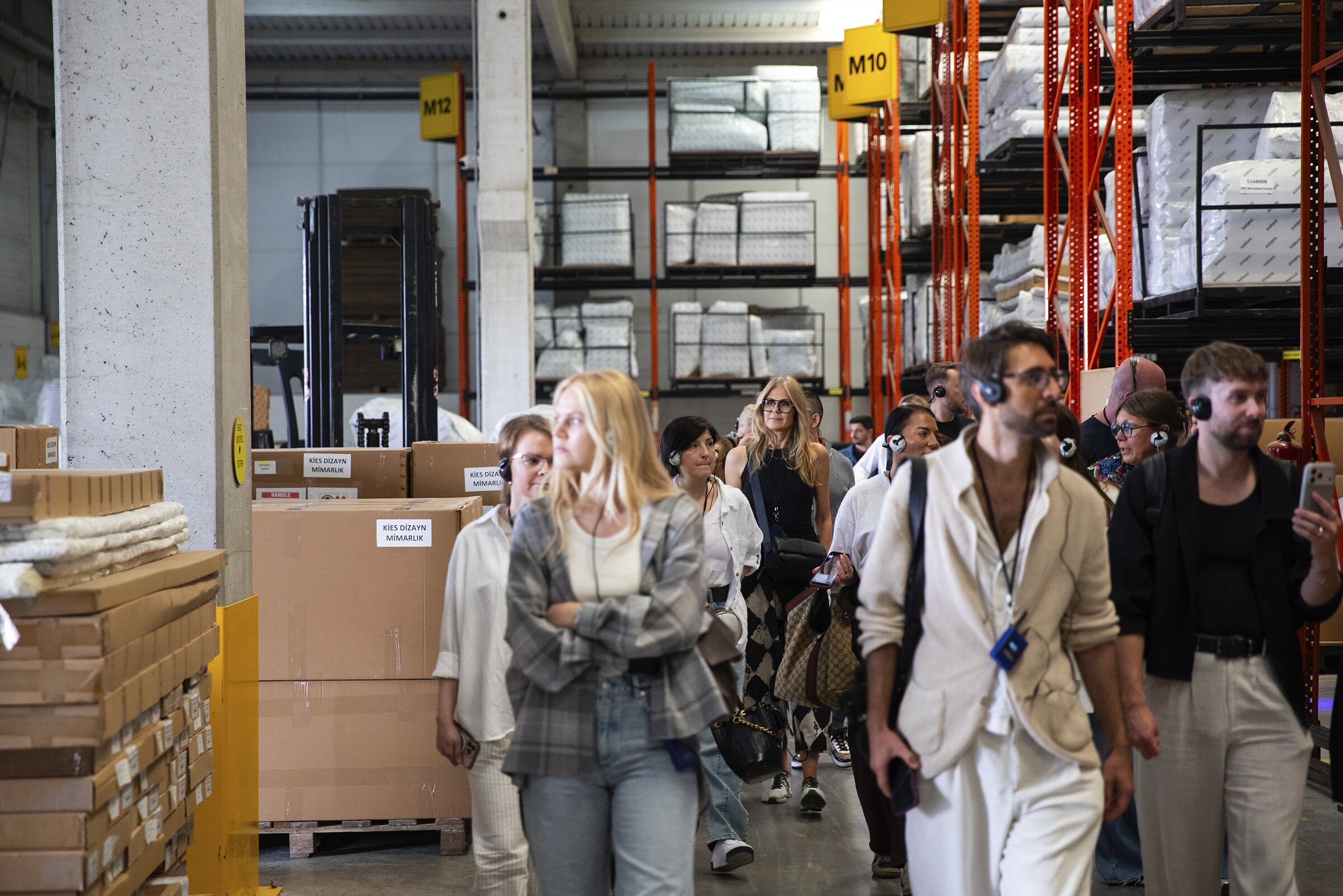 Busy warehouse with diverse group of people touring Saloni Furniture storage facility, showcasing organized shelving and furniture inventory.