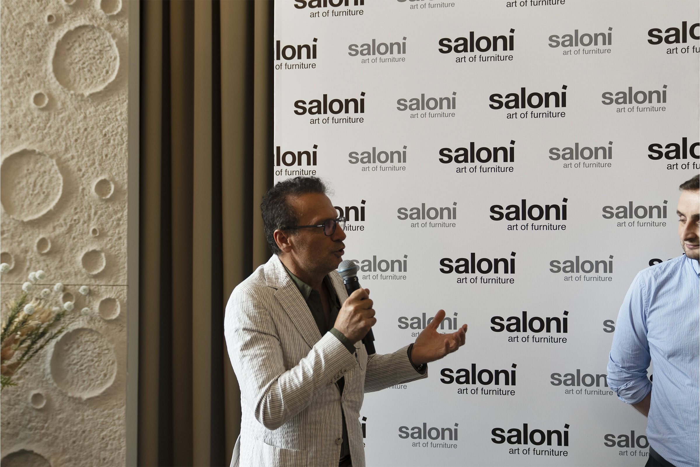 Modern man speaking at Saloni Furniture event with branded backdrop, highlighting contemporary furniture and interior design trends.
