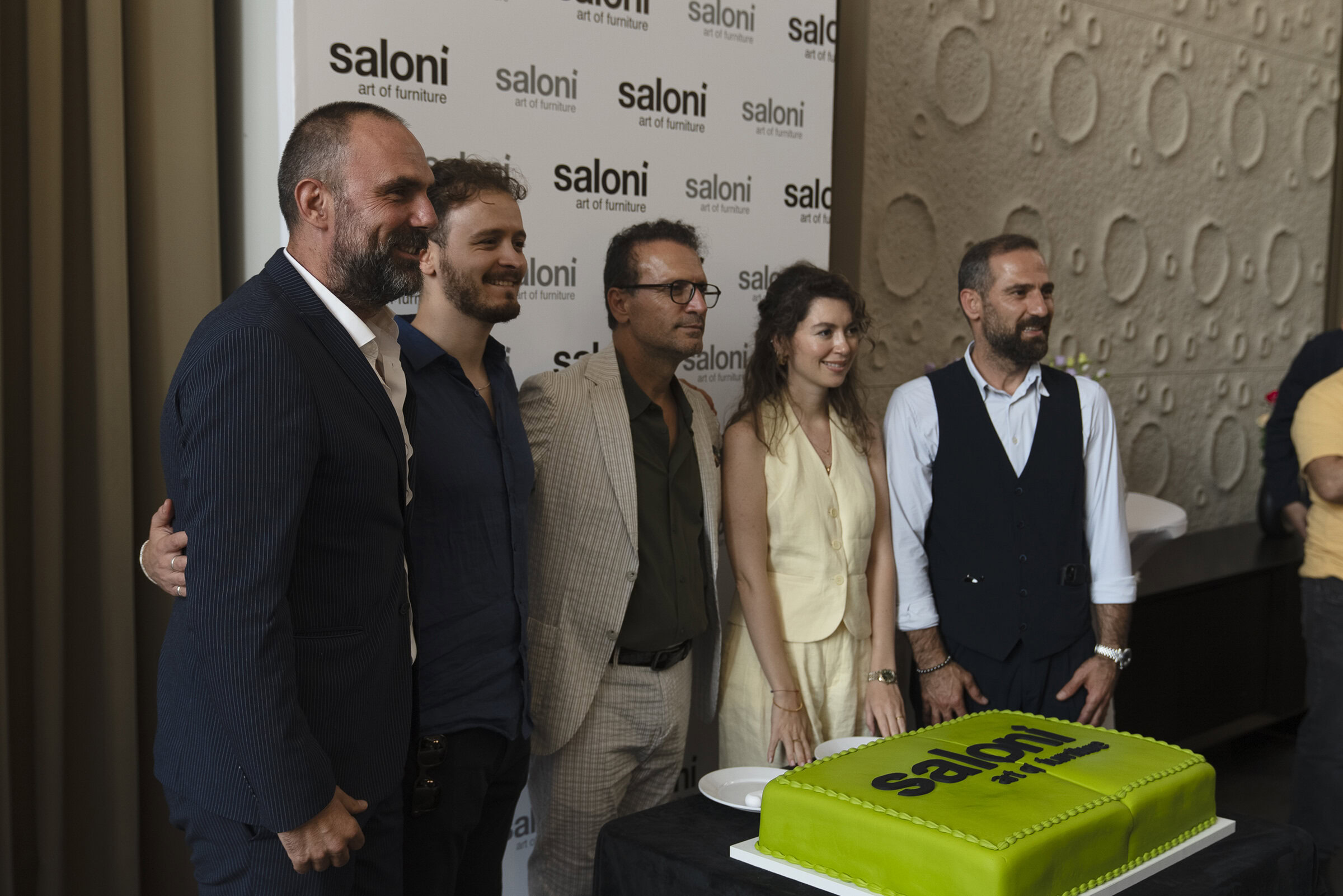 Elegant group of professionals celebrating at Saloni Furniture event, featuring modern furniture displays, with a bright green cake and stylish decor, highlighting luxury home furnishings and interior design.