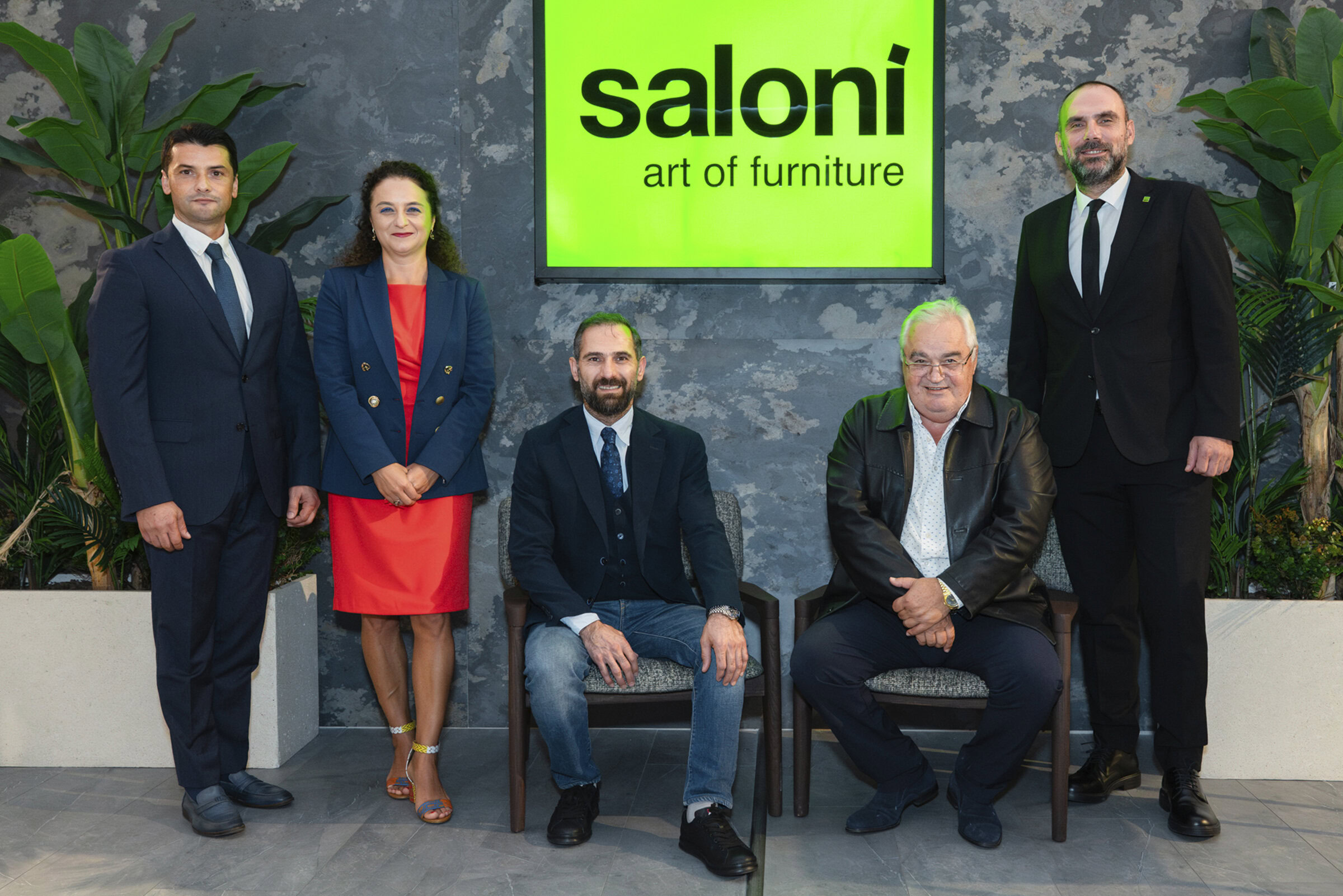 Modern furniture showroom team at Saloni Furniture with green logo background and indoor plants, showcasing professional staff and stylish ambiance.