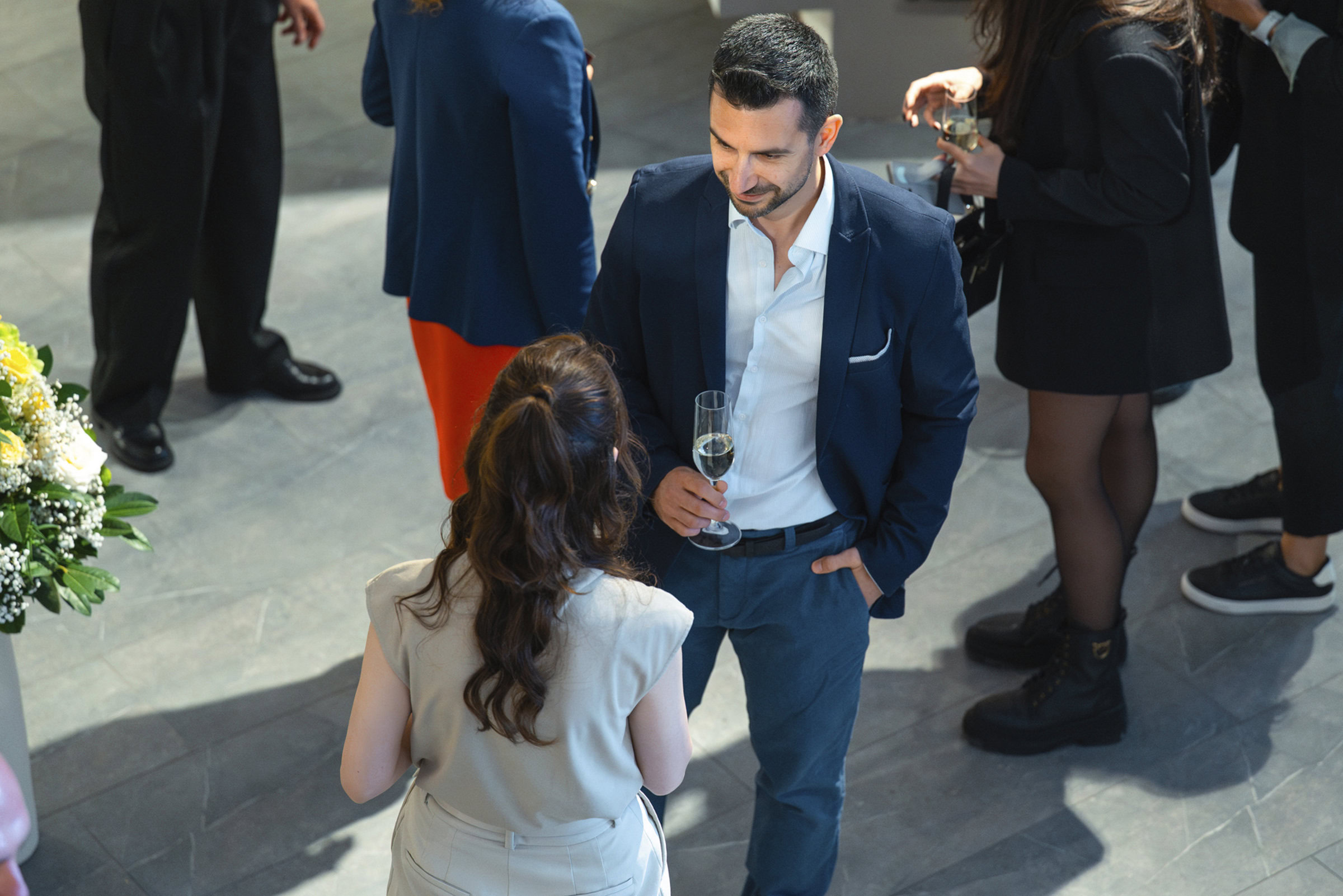 Elegant man and woman conversing at a stylish social event, holding champagne glasses, in a modern venue with contemporary furniture and floral decor, showcasing upscale lifestyle and event ambiance.