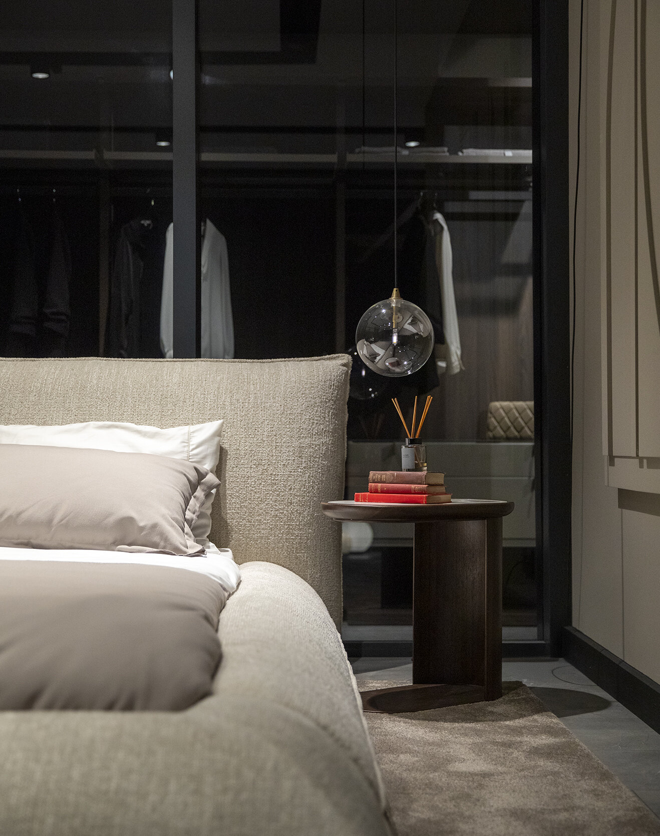 Luxurious bedroom with beige upholstered bed, neutral bedding, dark wooden side table with stacked books, modern hanging glass light fixture, and a sleek wardrobe in the background.